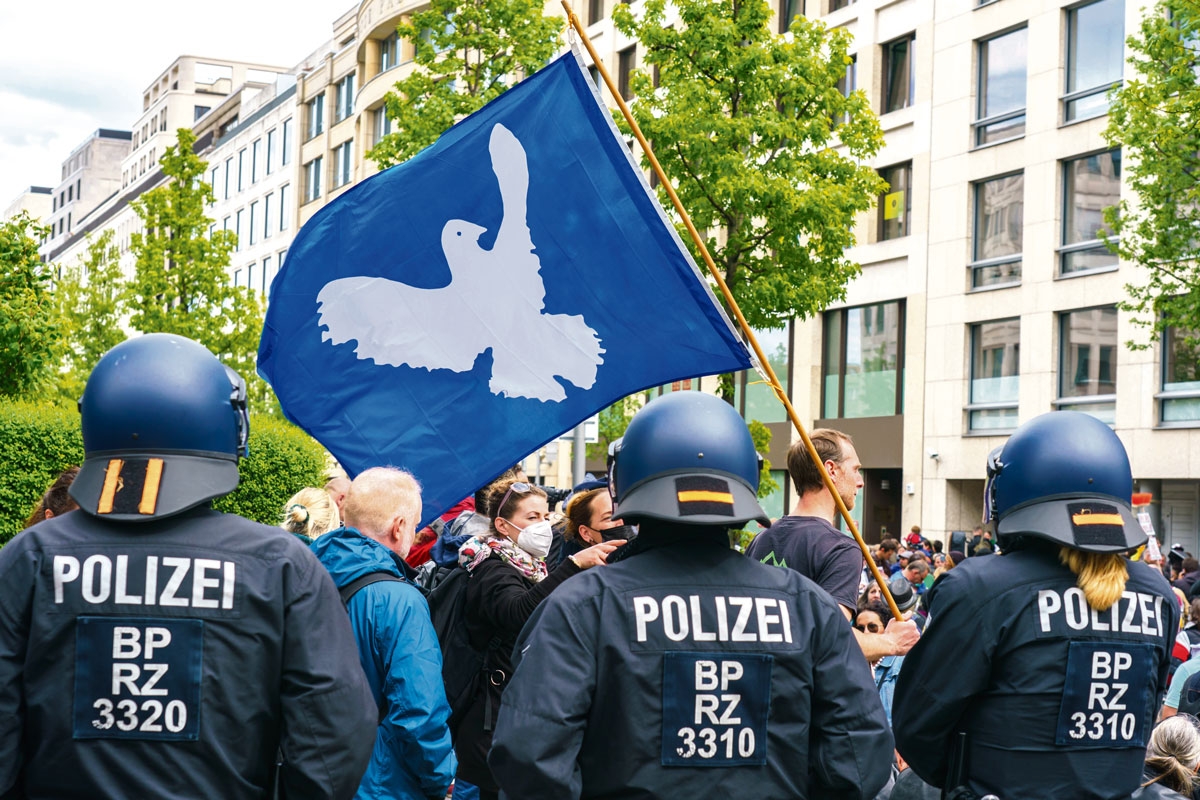 Hinter einer Reihe Polizisten stehen Demonstrant:innen, einer von ihnen trägt eine blaue Fahne mit Friedenstaube