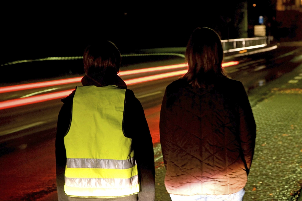 Zwei Personen stehen im Dunklen am Straßenrand, eine trägt eine Warnweste