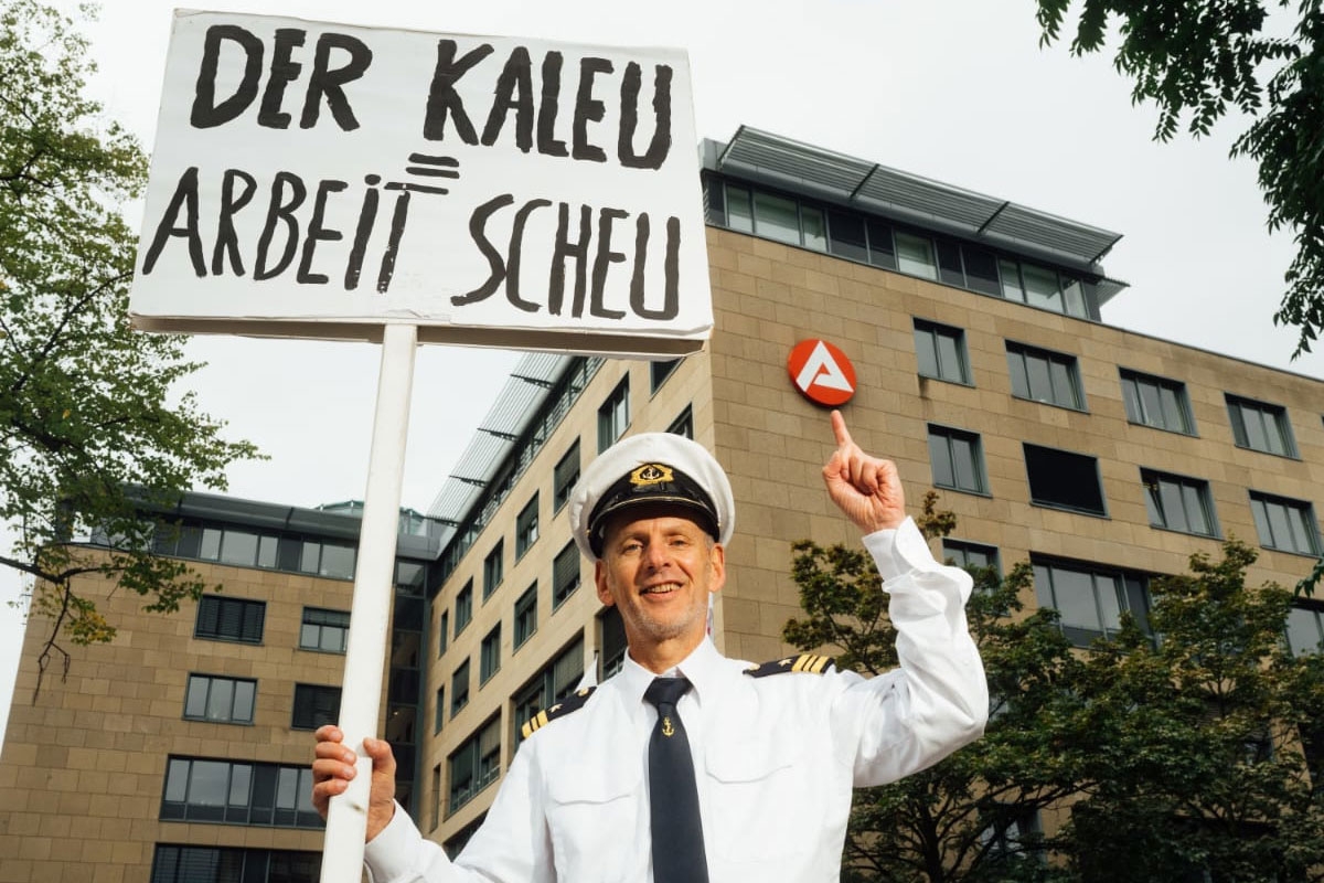 Der Kaleu mit einem Schild mit der Aufschrift »Der Kaleu = Arbeitsscheu«