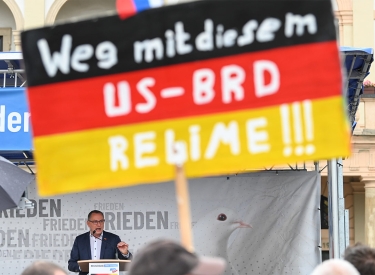 Schild in schwarz-rot-gold mit dem Spruch »Weg mit diesem US-BRD-Regime«, dahinter auf einer Bühne Chrupalla bei einer Rede