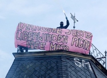 Vermummte Person auf Hausdach, daneben Transpi mit der Aufschrift: "Deutsche Justiz überwinden - ACAB - Free Maja"